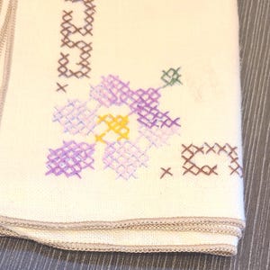 May include: Folded off-white cloth with embroidered floral and text details. The embroidery features purple, yellow, and green flowers, along with brown text and cross-stitch accents. The edges are finished with a light brown trim.