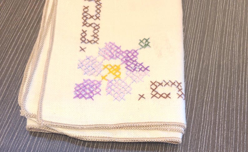 May include: Folded off-white cloth with embroidered floral and text details. The embroidery features purple, yellow, and green flowers, along with brown text and cross-stitch accents. The edges are finished with a light brown trim.