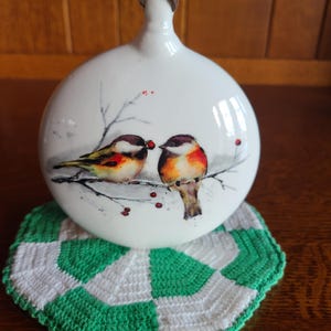 May include: A round white ceramic ornament featuring a painted illustration of two small birds perched on a thin branch with red berries. The birds have black heads, white cheeks, and orange-brown chests. The ornament has a small top loop.