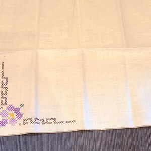 May include: White linen handkerchief with a cross-stitched floral design in purple, green, and yellow. The embroidery is in the corner of the handkerchief. The background is a dark wood surface.