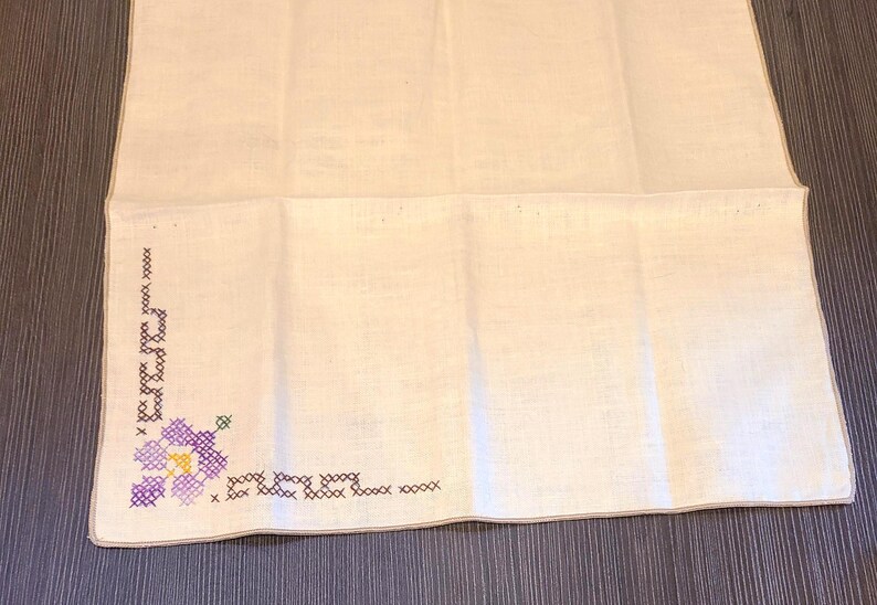 May include: White linen handkerchief with a cross-stitched floral design in purple, green, and yellow. The embroidery is in the corner of the handkerchief. The background is a dark wood surface.