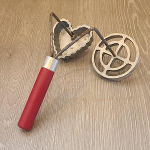 May include: A vintage cookie cutter with a red wooden handle, a heart-shaped cutter, and a round cutter. The metal cutters have a rustic, aged appearance, suggesting they are antique or well-used kitchen tools.