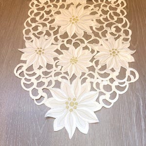 May include: Ivory-colored table runner with a floral poinsettia design. The runner features five large poinsettia flowers with gold-colored centers and detailed petal stitching. The edges are intricately cut with a delicate, openwork pattern.
