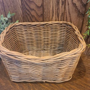 May include: A light brown woven wicker basket with a rectangular base and rounded corners. The basket features a thick, braided rim and a continuous, horizontal weave pattern throughout the sides, creating a sturdy, open-top storage container.