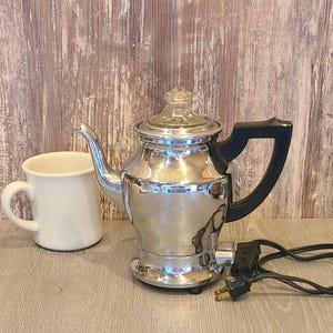 May include: A vintage chrome electric coffee pot with a black handle and a clear glass top. A white ceramic mug sits to the left of the coffee pot. The power cord is black.