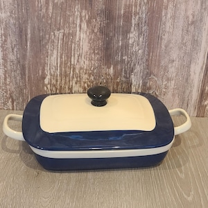 May include: A rectangular, ceramic baking dish with a lid. The dish is primarily navy blue with a cream-colored lid and white handles. The lid has a black knob. The dish is on a wooden surface.