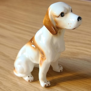 May include: A ceramic figurine of a seated dog. The dog has a white body with brown ears and patches. It has black eyes and a black nose. The figurine is on a wooden surface.