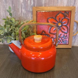 May include: A bright orange enamel tea kettle with a rounded body, a short spout, and a light-colored wooden handle. The lid features a matching light-colored wooden knob. The kettle has a smooth, glossy finish and a sturdy, functional design.