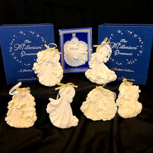 May include: A collection of white ceramic angel ornaments with gold ribbon hangers. The ornaments are in various poses and designs. The blue boxes feature the text "The Millennium Ornament" and "A Limited Edition".