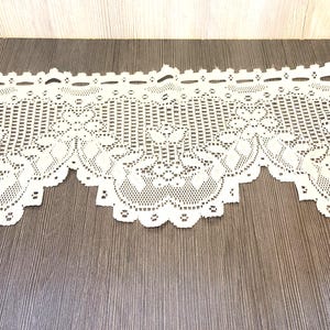 May include: Off-white lace trim with a scalloped edge. The lace features a detailed floral and geometric pattern. The trim is displayed on a dark wood-grain surface.