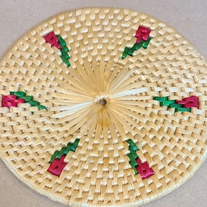 May include: A round, woven coaster with a natural beige color. The coaster features a repeating pattern of red and green embroidered flowers. The center of the coaster has a sunburst design.