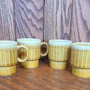 May include: Four vintage ceramic mugs with a stacked design. The mugs are a light yellow color with vertical ribbed detailing and a cream-colored rim. The mugs have a handle and are sitting on a wooden surface.