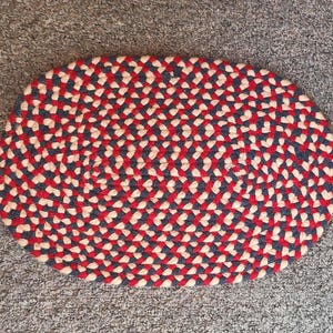 May include: A round braided rug with a red, white, and blue pattern. The rug is made of thick, woven yarn and has a slightly textured surface.
