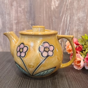 May include: A brown ceramic teapot with a floral design. The teapot has two pink flowers with black outlines and green leaves. The teapot has a lid and a handle.