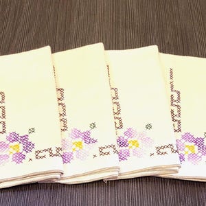 May include: Four folded cream napkins with embroidered floral patterns. The napkins display purple and yellow flowers with brown and green details. They are stacked on a dark brown wooden surface.
