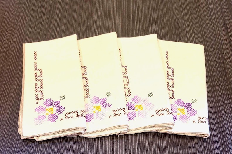 May include: Four folded cream napkins with embroidered floral patterns. The napkins display purple and yellow flowers with brown and green details. They are stacked on a dark brown wooden surface.