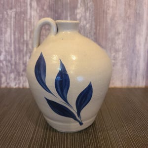 May include: A cream-colored ceramic jug with a single handle and a narrow neck. The body of the jug features a hand-painted design of three dark blue leaves extending from a single stem, with a smooth, glossy finish over the entire surface.
