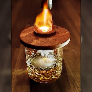 Op de afbeelding: Een helder glas met een houten deksel wordt met een brander aangestoken. Het glas bevat een heldere vloeistof en een witte rookwolk. Het glas staat op een houten oppervlak.