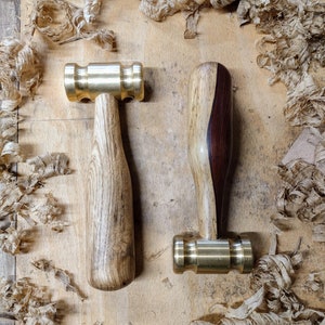 May include: Two brass hammers with wooden handles. The hammers are on a wooden surface with wood shavings.