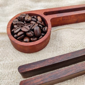 May include: A wooden scoop filled with dark brown coffee beans, resting on a beige fabric surface. The scoop is part of a larger wooden serving tray with a second, smaller scoop.