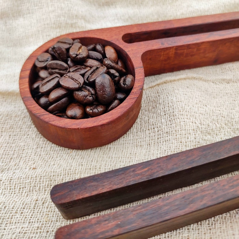 May include: A wooden scoop filled with dark brown coffee beans, resting on a beige fabric surface. The scoop is part of a larger wooden serving tray with a second, smaller scoop.