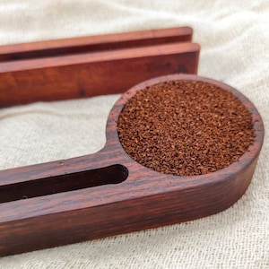May include: A wooden coffee scoop with a dark brown finish. The scoop has a small compartment for holding coffee grounds and a larger compartment for holding coffee beans. The scoop is filled with coffee grounds.