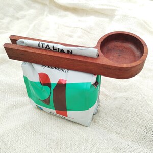 May include: A wooden scoop with a handle and a round bowl, used for scooping coffee beans. The scoop is resting on top of a bag of "Italian" coffee beans by Sainsbury's.