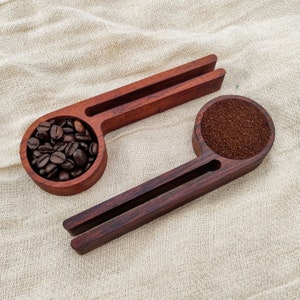 May include: Two wooden scoops with a handle. One scoop is filled with coffee beans, the other with ground coffee. The scoops are on a beige fabric background.