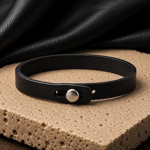 May include: A simple black leather bracelet featuring a smooth, matte finish and a single circular silver-toned metal stud closure. The band is constructed from a flat strip of leather that loops around to secure with the metal fastener at the front.