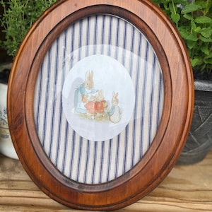 May include: Oval wooden picture frame with a watercolor illustration of rabbits. The frame has a dark brown finish and a glass cover. The background features blue and white striped fabric.