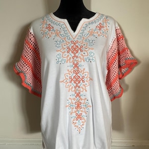 May include: A white short-sleeved top featuring a V-neckline with intricate coral and light blue embroidery down the center. The sleeves are made of a coral-colored crochet knit fabric with a scalloped edge and light blue trim accents.