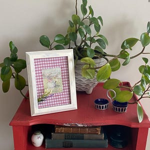 May include: A white wooden picture frame containing a red and white gingham background with a small illustration of a figure in a landscape, accented by a sprig of dried lavender. The frame sits on a red table with books and small containers.