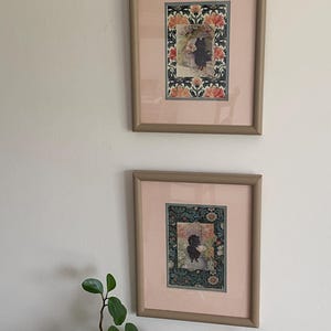May include: Two framed rectangular prints featuring black cat illustrations centered within floral borders. Each print is encased in a light pink mat and a smooth, neutral-toned frame. The artwork displays intricate botanical patterns with orange and green accents.