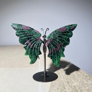 May include: A butterfly-shaped decorative object carved from green zoisite with embedded magenta ruby inclusions. The wings are mounted on a thin metal rod attached to a circular black base, featuring two small wire antennae at the top centre.