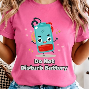 Pode incluir: Uma t-shirt rosa brilhante com um gráfico de desenho animado de uma bateria turquesa com uma secção vermelha de carga baixa, uma cara feliz e braços levantados. Abaixo do gráfico, o texto "Do Not Disturb Battery" em letras pretas.
