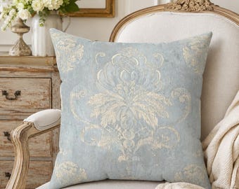 May include: A square throw pillow featuring a light blue fabric base with a distressed, cream-colored damask pattern. The design displays symmetrical, ornate floral and scrollwork motifs centered on the front, with a textured, weathered appearance.