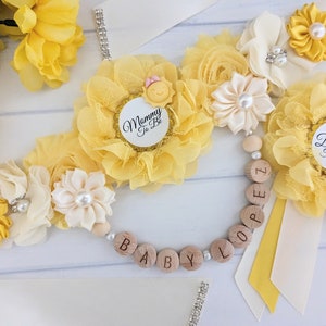May include: Yellow and white flower sashes with the text "Mommy to Be" and "Daddy to Be" on them. A wooden bead necklace with the word "BABY" spelled out on it.