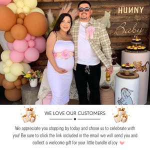 May include: A pregnant woman and a man stand in front of a wooden sign that says "Hunny Baby". The woman is wearing a white dress with a pink flower sash. The man is wearing a plaid shirt and a pink ribbon. There are balloons and decorations in the background.