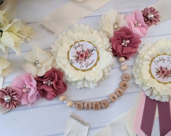 May include: A white satin sash with pink and burgundy fabric flowers, a wooden bead necklace with the words "BABY NAME" and two white fabric flowers with gold glitter centers that say "Baby In Bloom" and "Daddy To Be".
