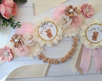 May include: A pink and cream floral sash with a gold glitter 'Mommy To Be' and 'Daddy To Be' pin with a Highland Cow design. The sash also includes a wooden bead necklace with the word 'Baby' spelled out.