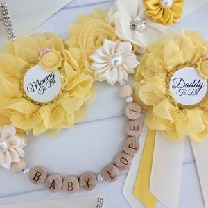 May include: Yellow and white flower corsages with a sun and the text "Mommy to Be" and "Daddy to Be" on the center of each corsage. A wooden bead necklace with the letters "BABY LOOP" is included.