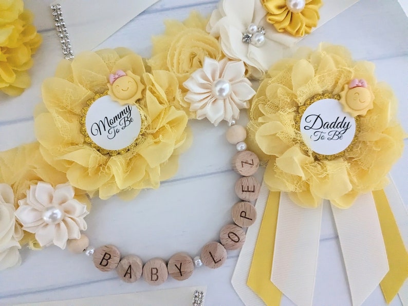May include: Yellow and white flower corsages with a sun and the text "Mommy to Be" and "Daddy to Be" on the center of each corsage. A wooden bead necklace with the letters "BABY LOOP" is included.