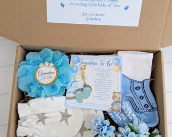 May include: A baby shower gift box with blue and white items. The box contains a blue flower, baby socks, mittens, and a charm with baby feet. The box also includes a card that says "Grandma To Be".