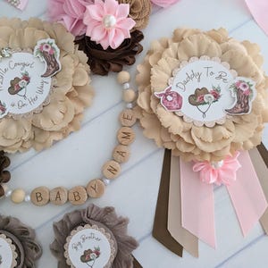 May include: A set of six brown and pink flower pins with ribbons. The pins feature a variety of designs, including a cowboy hat, a horseshoe, and the words "A Little Cowgirl is on Her Way", "Daddy to Be", "Big Sister", and "Big Brother". A wooden bead necklace with the words "BABY" and "NAME" is also included.