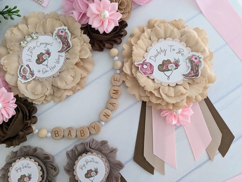 May include: A set of six brown and pink flower pins with ribbons. The pins feature a variety of designs, including a cowboy hat, a horseshoe, and the words "A Little Cowgirl is on Her Way", "Daddy to Be", "Big Sister", and "Big Brother". A wooden bead necklace with the words "BABY" and "NAME" is also included.