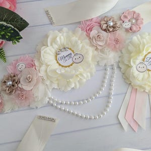 May include: A white satin sash with a pearl necklace and pink and white flowers. The sash has the text "Mommy to Be" and "Daddy to Be" on it.