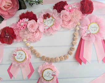 May include: Pink and red flower crown with a wooden bead garland that says "Our Cherry On Top". The garland is decorated with small white beads. There are also two smaller pink flower pins that say "Big Sister" and "Big Brother".