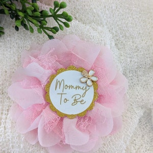 May include: Pink and white flower with a gold glitter circle that says "Mommy To Be" in white text. The flower has a white flower with a gold center on top.