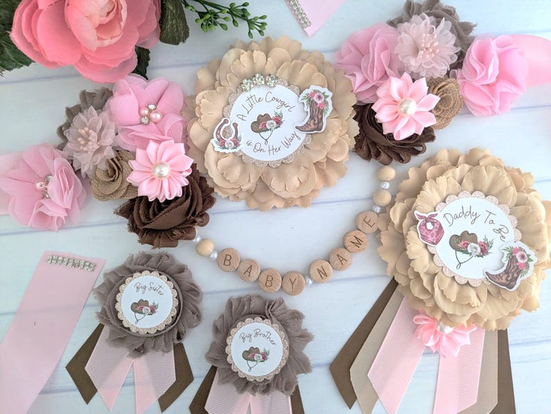 May include: A set of six brown and pink flower sashes with a cowboy theme. The sashes feature the text "A Little Cowgirl on Her Way", "Big Sister", "Big Brother", "Daddy to Be", and "Baby Name".