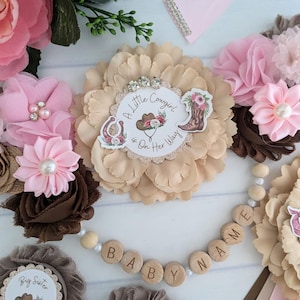 May include: A pink and brown baby shower decoration with a flower garland and a wooden bead garland that says "Baby Name". The flower garland has a brown flower with a white circle that says "A Little Cowgirl is On Her Way".